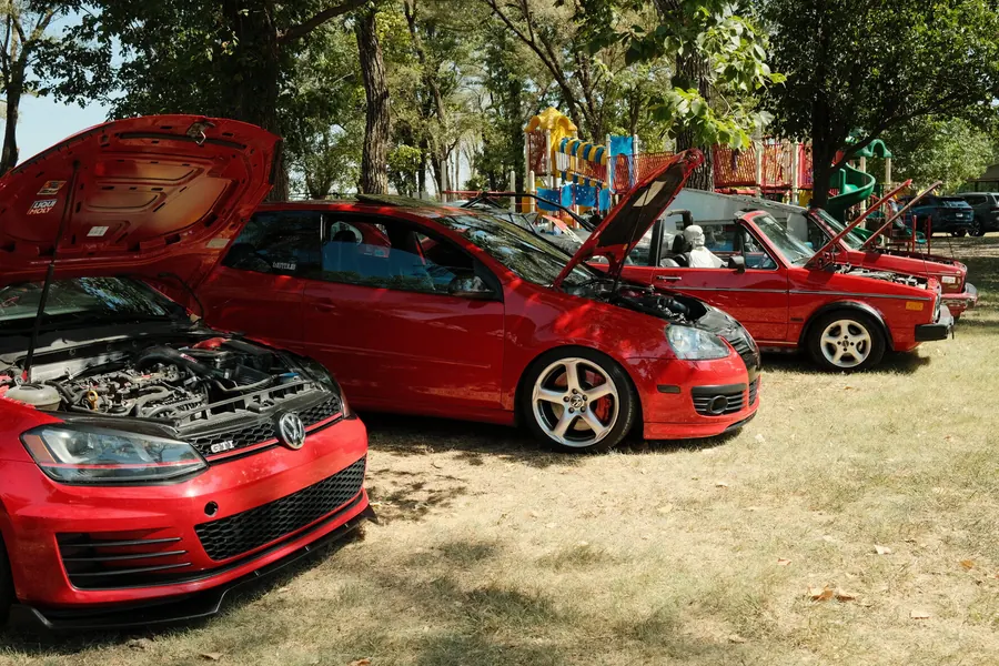 A row of red Volkswagens with their hoods up on the grass at the 2026 show: two GTIs in front and two older convertibles behind